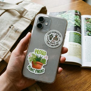May include: A clear smartphone case decorated with two stickers. One circular sticker features a stack of books, a potted plant, and a cup with the text EASILY DISTRACTED BY PLANTS AND BOOKS. The other sticker shows a potted succulent with the text PROUD PLANT PARENT.