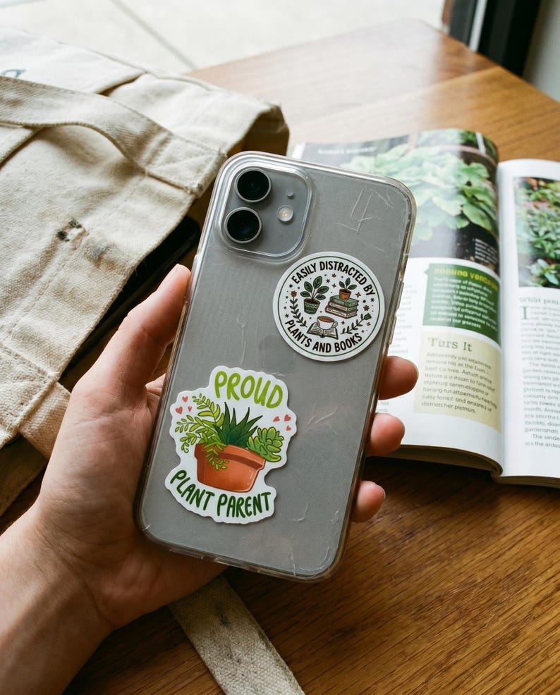 May include: A clear smartphone case decorated with two stickers. One circular sticker features a stack of books, a potted plant, and a cup with the text EASILY DISTRACTED BY PLANTS AND BOOKS. The other sticker shows a potted succulent with the text PROUD PLANT PARENT.
