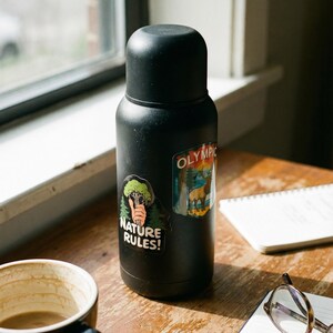 May include: A black insulated water bottle with two stickers, one reading "NATURE RULES!" and the other with "OLYMPIC" text. The bottle sits on a wooden surface next to a coffee mug, notebook, and glasses.