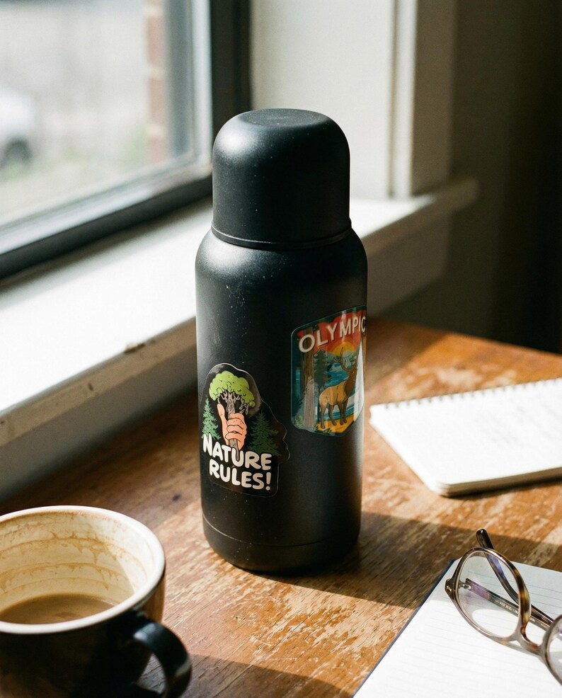 May include: A black insulated water bottle with two stickers, one reading "NATURE RULES!" and the other with "OLYMPIC" text. The bottle sits on a wooden surface next to a coffee mug, notebook, and glasses.