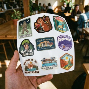 May include: A sheet of colorful stickers featuring nature-themed designs. The stickers include images of Bigfoot, a squirrel, mountains, a camper van, and the text "Nature Rules!" and "Grand Canyon".
