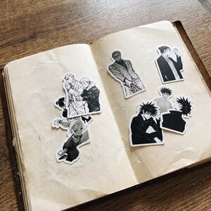 May include: A collection of black and white anime-style stickers featuring various characters, including figures in suits and casual attire, arranged on the open pages of a notebook with cream-coloured paper and a visible texture.