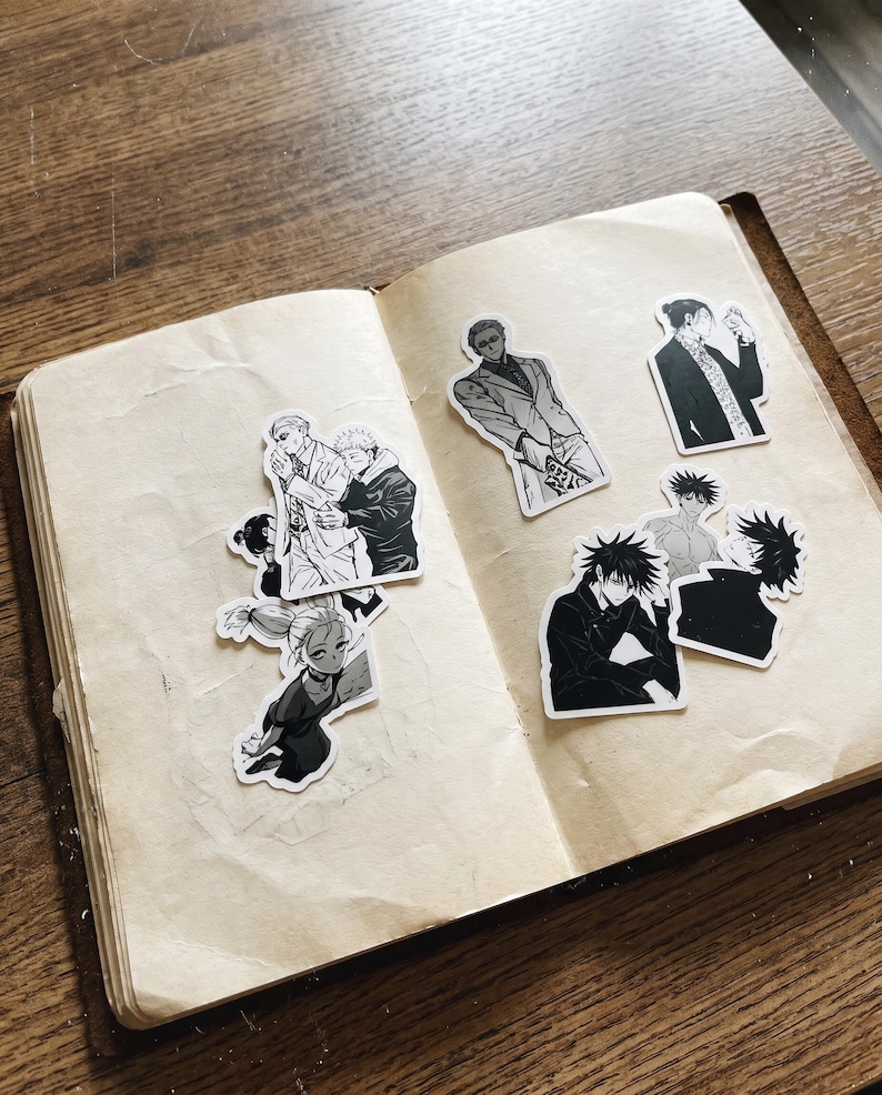 May include: A collection of black and white anime-style stickers featuring various characters, including figures in suits and casual attire, arranged on the open pages of a notebook with cream-coloured paper and a visible texture.