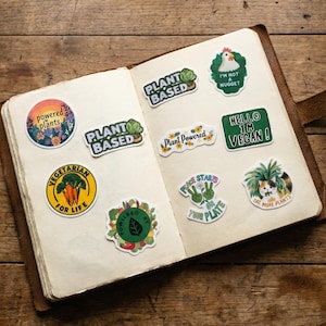 May include: A spread of colorful stickers with plant-based and vegan themes. The stickers feature text like "Plant Based", "Vegetarian for Life", and "Hello I'm Vegan!" on a wooden background.