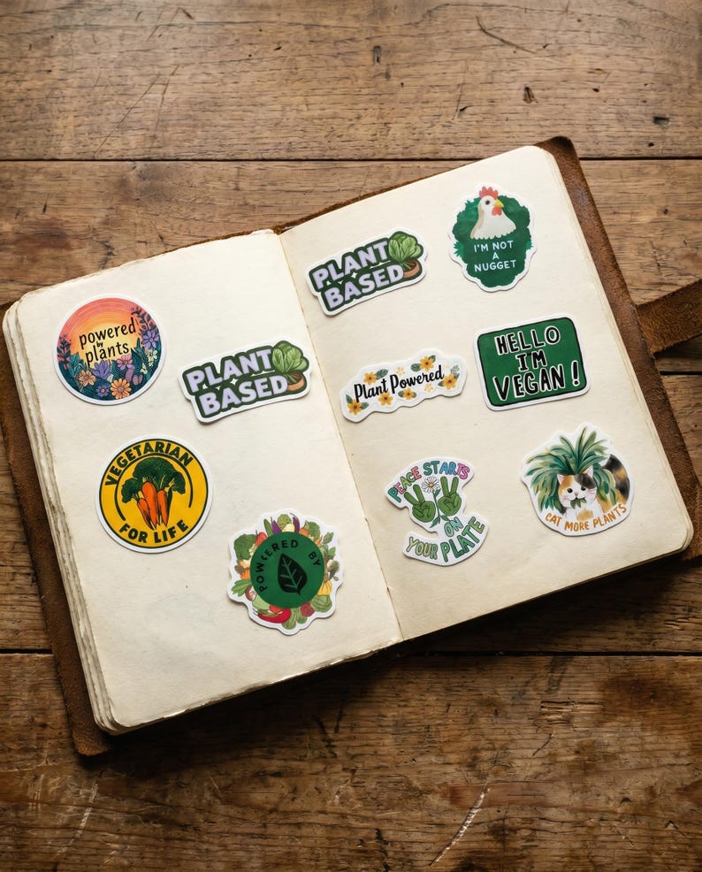 May include: A spread of colorful stickers with plant-based and vegan themes. The stickers feature text like "Plant Based", "Vegetarian for Life", and "Hello I'm Vegan!" on a wooden background.