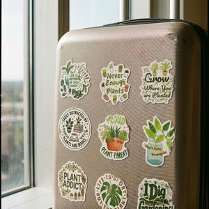 May include: A beige hard-shell suitcase decorated with nine plant-themed stickers featuring various green foliage illustrations and text including Life is Better in the Garden, Never Enough Plants, Grow Where You are Planted, and I Dig Gardening.