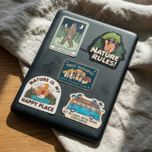 May include: A black laptop covered in colorful nature-themed stickers. Stickers include images of Bigfoot, a mountain scene, mushrooms, and a blue and orange van with the text "Get in Loser, We're Going Everywhere."