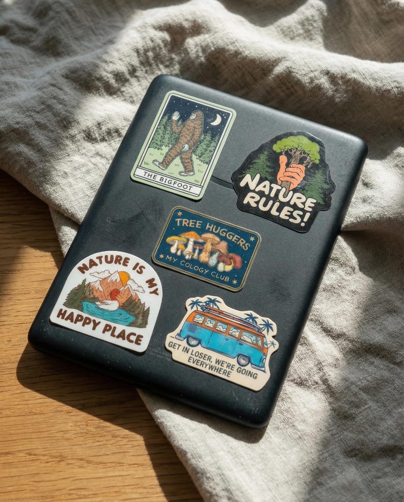 May include: A black laptop covered in colorful nature-themed stickers. Stickers include images of Bigfoot, a mountain scene, mushrooms, and a blue and orange van with the text "Get in Loser, We're Going Everywhere."