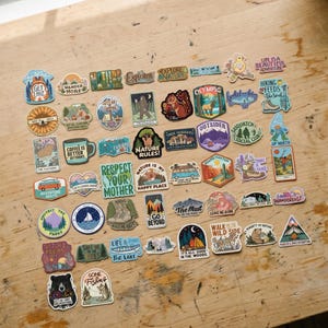 May include: A collection of colorful outdoor-themed stickers with various designs and text. The stickers feature images of mountains, forests, animals, and phrases like "Explore Nature" and "Wander More". They are arranged on a wooden surface.