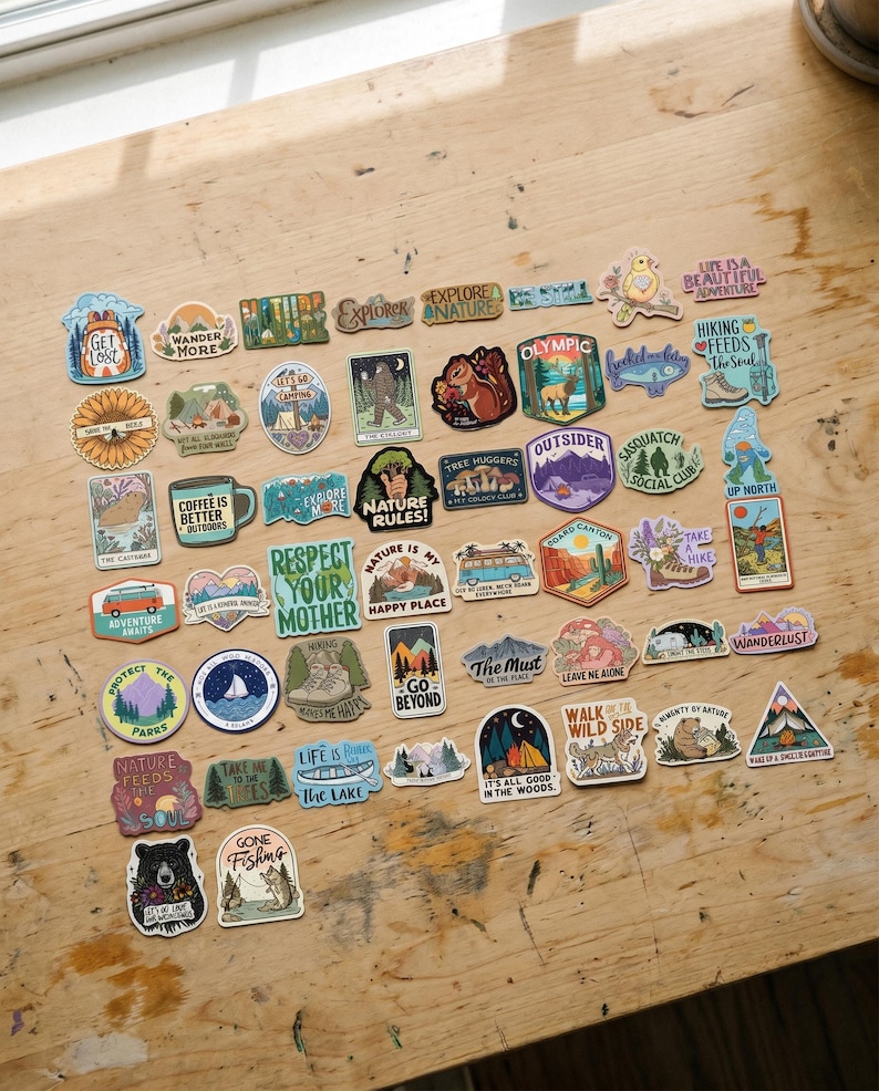 May include: A collection of colorful outdoor-themed stickers with various designs and text. The stickers feature images of mountains, forests, animals, and phrases like "Explore Nature" and "Wander More". They are arranged on a wooden surface.