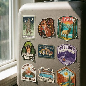May include: A collection of colorful, nature-themed stickers adhered to a gray suitcase. Designs include a Bigfoot, a squirrel, an elk in Olympic National Park, and phrases like "Nature Rules!" and "Happy Place."