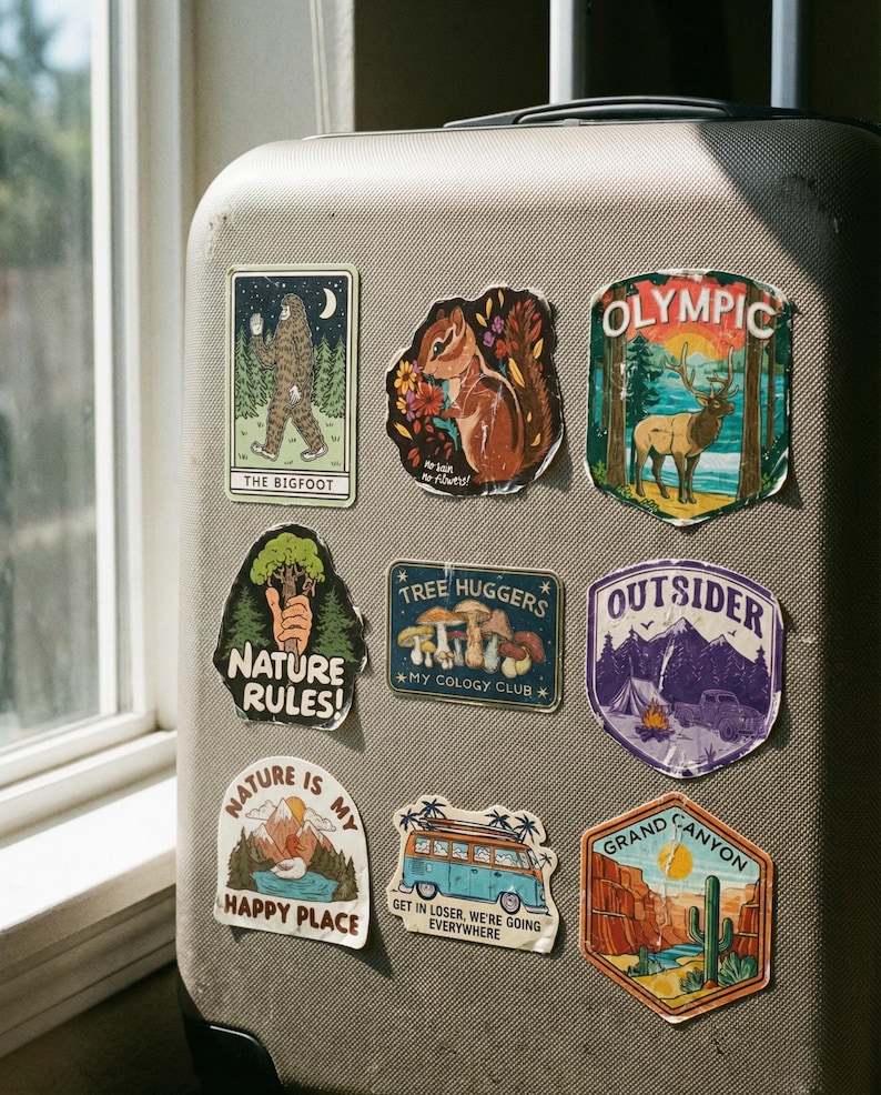 May include: A collection of colorful, nature-themed stickers adhered to a gray suitcase. Designs include a Bigfoot, a squirrel, an elk in Olympic National Park, and phrases like "Nature Rules!" and "Happy Place."