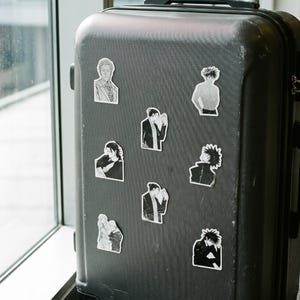 May include: A dark grey hard-shell rolling suitcase decorated with eight individual black and white anime-style character stickers. The stickers feature various figures in different poses, including portraits and upper-body illustrations, arranged across the front surface of the luggage.
