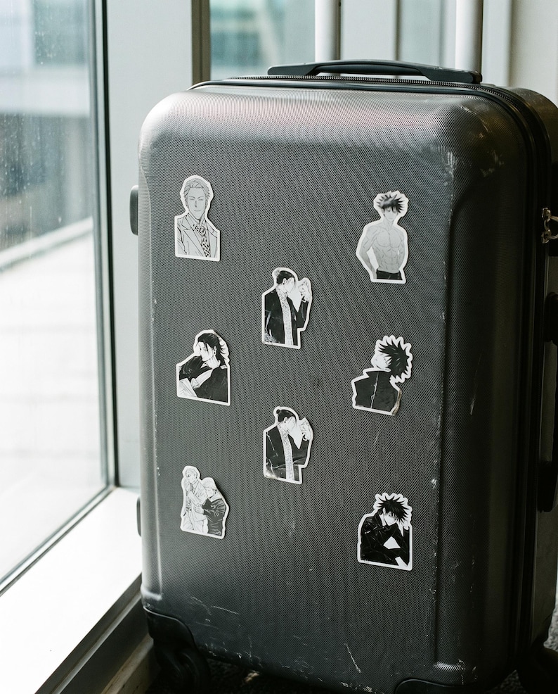 May include: A dark grey hard-shell rolling suitcase decorated with eight individual black and white anime-style character stickers. The stickers feature various figures in different poses, including portraits and upper-body illustrations, arranged across the front surface of the luggage.