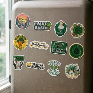 May include: A collection of colorful vegan-themed stickers on a tan suitcase. The stickers feature phrases like "Plant Based," "Hello I'm Vegan!", and "Eat More Plants." Other stickers have illustrations of vegetables and animals.