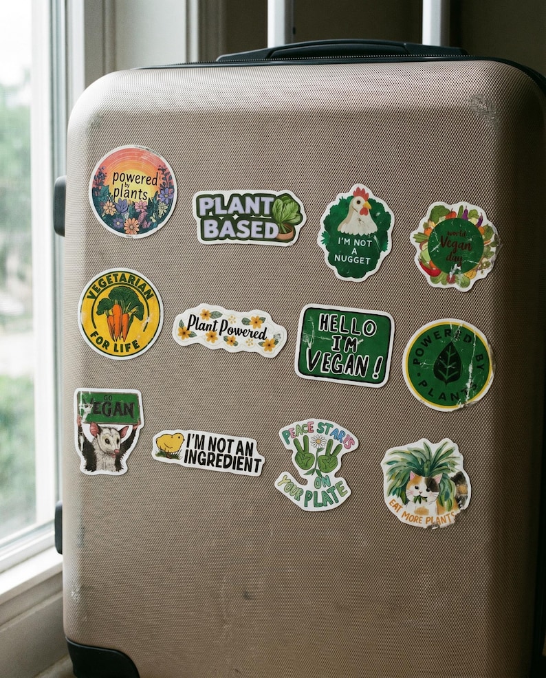 May include: A collection of colorful vegan-themed stickers on a tan suitcase. The stickers feature phrases like "Plant Based," "Hello I'm Vegan!", and "Eat More Plants." Other stickers have illustrations of vegetables and animals.