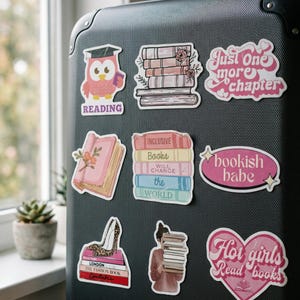 May include: A collection of colorful stickers on a black suitcase. The stickers feature book-related designs, including stacks of books, an owl with a graduation cap, and phrases like "Just One More Chapter" and "Hot girls Read books".
