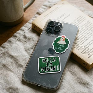 May include: A clear phone case with two stickers. One sticker says "I'M NOT A NUGGET" with a cartoon chicken. The other sticker says "HELLO I'M VEGAN!" in white text on a green background. A coffee mug and a book are in the background.