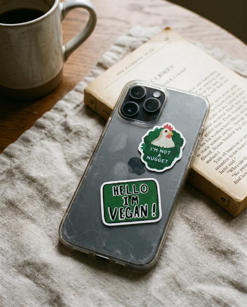 May include: A clear phone case with two stickers. One sticker says "I'M NOT A NUGGET" with a cartoon chicken. The other sticker says "HELLO I'M VEGAN!" in white text on a green background. A coffee mug and a book are in the background.