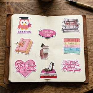 May include: An open notebook with various stickers featuring book-related themes. Stickers include an owl with a graduation cap, stacks of books, and phrases like "Hot girls Read books" and "Just one more chapter."