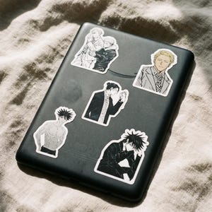 May include: A black rectangular electronic device decorated with five distinct anime-style stickers. The stickers feature various characters in different poses, including a shirtless figure, a character in a suit, and others in dark clothing.