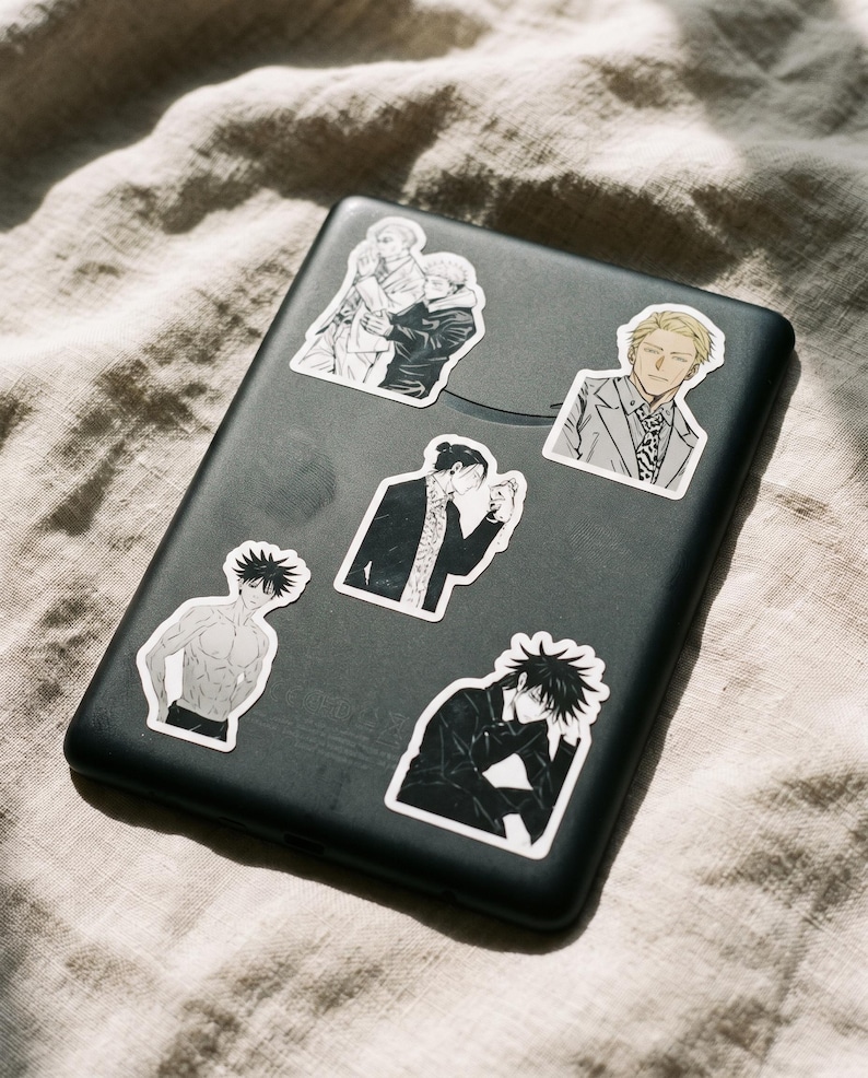 May include: A black rectangular electronic device decorated with five distinct anime-style stickers. The stickers feature various characters in different poses, including a shirtless figure, a character in a suit, and others in dark clothing.