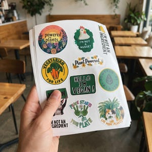 May include: A stack of stickers with various designs and text, including "Powered Plants," "I'm Not a Nugget," and "Vegetarian for Life." The stickers feature colorful illustrations of plants, animals, and flowers, promoting vegan and plant-based lifestyles.