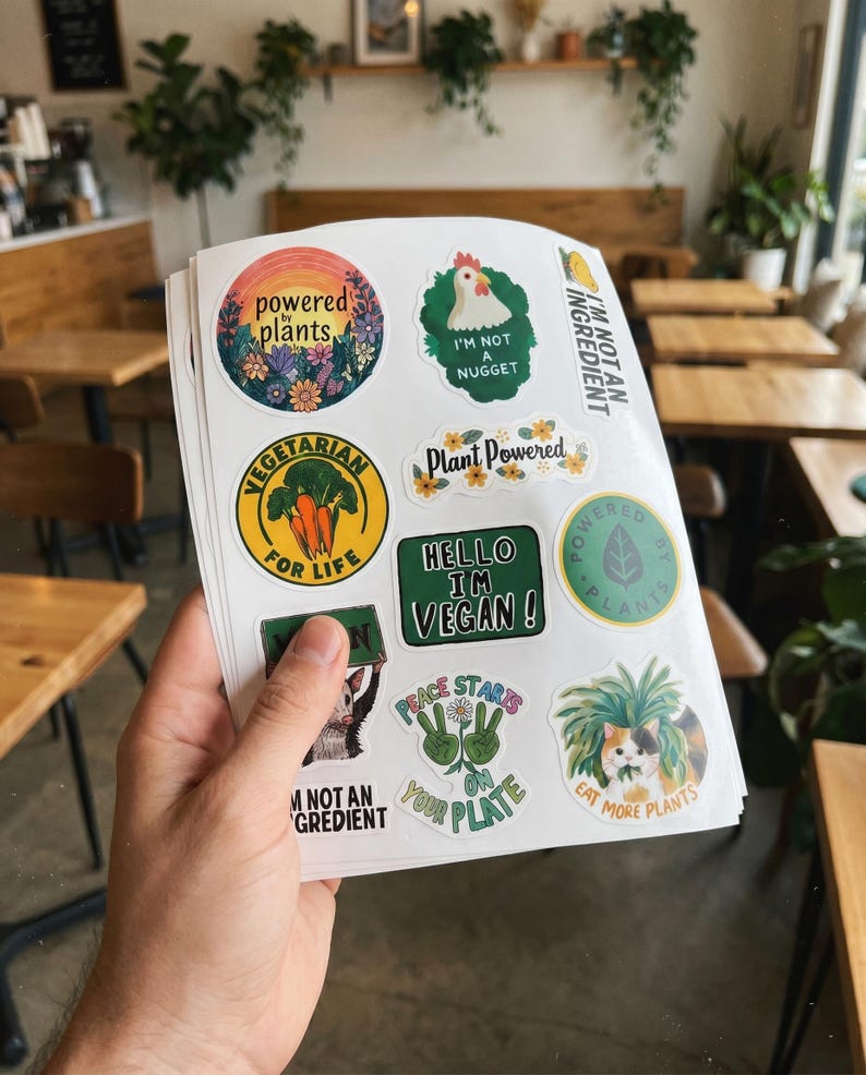 May include: A stack of stickers with various designs and text, including "Powered Plants," "I'm Not a Nugget," and "Vegetarian for Life." The stickers feature colorful illustrations of plants, animals, and flowers, promoting vegan and plant-based lifestyles.