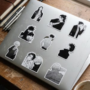 May include: A silver laptop lid decorated with nine individual black and white anime-style stickers. The stickers feature various characters in different poses, including portraits, a shirtless figure, and a pair of characters making a peace sign.