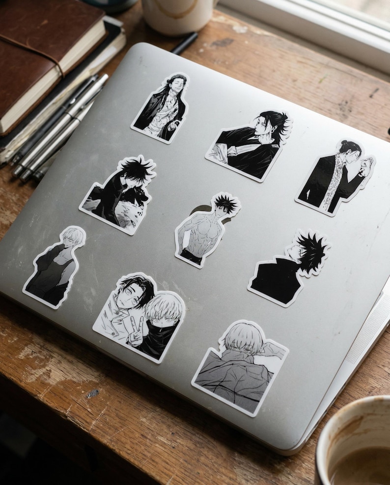May include: A silver laptop lid decorated with nine individual black and white anime-style stickers. The stickers feature various characters in different poses, including portraits, a shirtless figure, and a pair of characters making a peace sign.