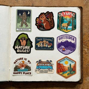May include: A collection of colorful stickers with nature-themed designs. The stickers feature images of Bigfoot, a chipmunk, a deer, mountains, a camper, a Grand Canyon scene, and text that reads "Nature Rules!" and "Nature is my happy place."