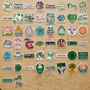 May include: A collection of colorful vegan-themed stickers with various designs and text. The stickers feature phrases like "Vegan Vibes," "Powered by Plants," and "Eat Plants." The stickers are arranged on a wooden surface.