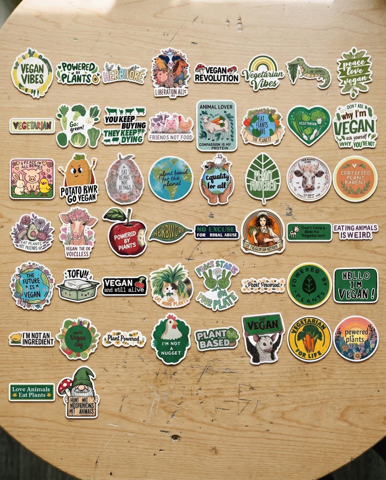 May include: A collection of colorful vegan-themed stickers with various designs and text. The stickers feature phrases like "Vegan Vibes," "Powered by Plants," and "Eat Plants." The stickers are arranged on a wooden surface.