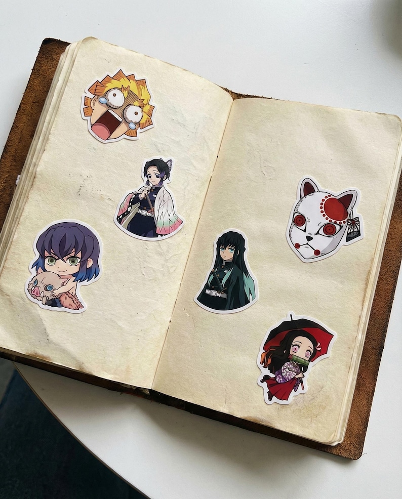 May include: A selection of anime character stickers, featuring a character with yellow hair and a surprised expression, a character with a fox mask, and more. The stickers are arranged on the open pages of a vintage-style journal.