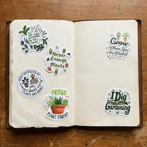 May include: Five decorative stickers with plant-themed illustrations and text on the pages of an open notebook. The stickers feature phrases including Life is Better in the Garden, Never Enough Plants, Easily Distracted by Plants and Books, Grow Where You are Planted, Proud Plant Parent, and I Dig Gardening.
