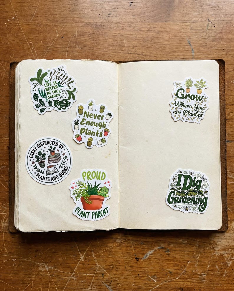 May include: Five decorative stickers with plant-themed illustrations and text on the pages of an open notebook. The stickers feature phrases including Life is Better in the Garden, Never Enough Plants, Easily Distracted by Plants and Books, Grow Where You are Planted, Proud Plant Parent, and I Dig Gardening.