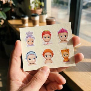 May include: A sheet of stickers featuring six small doll-like figures with various head coverings. The stickers have a cream-coloured background and are held in a person's hand. The figures have large eyes and are in different colours.