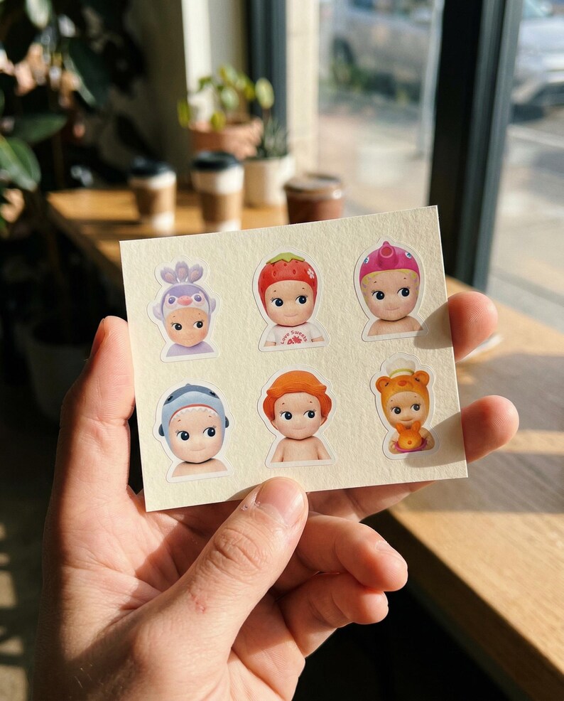 May include: A sheet of stickers featuring six small doll-like figures with various head coverings. The stickers have a cream-coloured background and are held in a person's hand. The figures have large eyes and are in different colours.