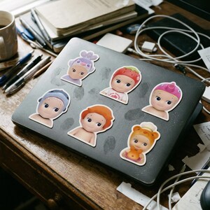 May include: A selection of vinyl stickers depicting baby-faced characters with different headwear, such as a strawberry, a bear, and a lavender bonnet. The stickers are arranged on a grey laptop, alongside pens, notebooks, and other desk accessories.