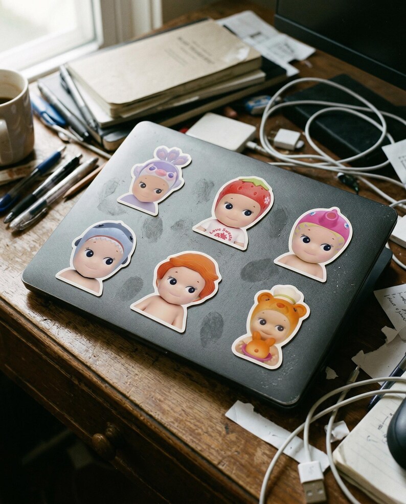 May include: A selection of vinyl stickers depicting baby-faced characters with different headwear, such as a strawberry, a bear, and a lavender bonnet. The stickers are arranged on a grey laptop, alongside pens, notebooks, and other desk accessories.