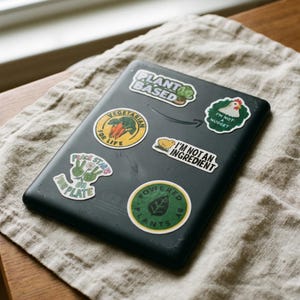 May include: A black electronic device covered in colorful stickers with text like "Plant Based", "Vegetarian for Life", and "Powered by Plants". The stickers feature images of vegetables, leaves, and a chicken. The device rests on a beige linen cloth.