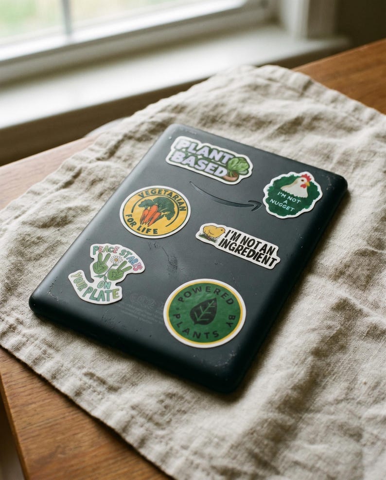 May include: A black electronic device covered in colorful stickers with text like "Plant Based", "Vegetarian for Life", and "Powered by Plants". The stickers feature images of vegetables, leaves, and a chicken. The device rests on a beige linen cloth.