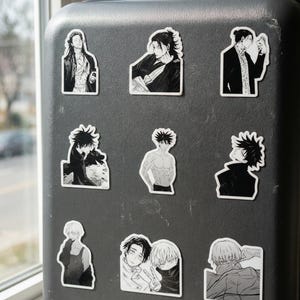 May include: A black suitcase featuring nine individual black and white vinyl stickers. The stickers display various anime-style character illustrations, including portraits, figures in dark clothing, and scenes with multiple characters in a manga art style.