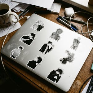 May include: A silver laptop lid decorated with nine distinct black and white anime-style character stickers. The stickers feature various figures in different poses, including portraits and full-body illustrations, arranged across the metallic surface.
