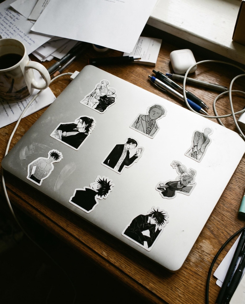 May include: A silver laptop lid decorated with nine distinct black and white anime-style character stickers. The stickers feature various figures in different poses, including portraits and full-body illustrations, arranged across the metallic surface.