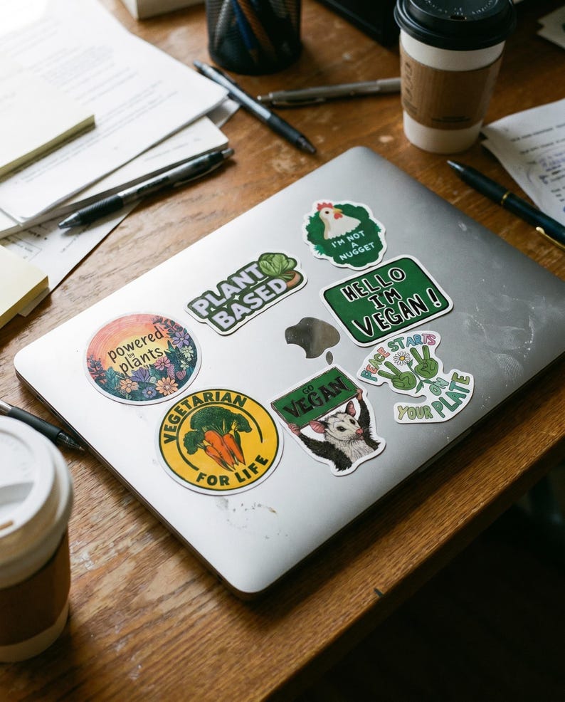 May include: A silver laptop covered in colorful stickers with vegan and vegetarian themes. Stickers include phrases like "Plant Based", "Hello I'm Vegan", and "Vegetarian for Life". The laptop sits on a wooden desk with pens and papers.