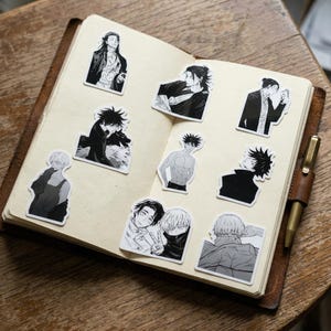 May include: An open notebook with a leather cover containing nine black and white stickers featuring various anime-style character illustrations. The stickers depict different figures in manga art style, including portraits and action poses, arranged on the pages.