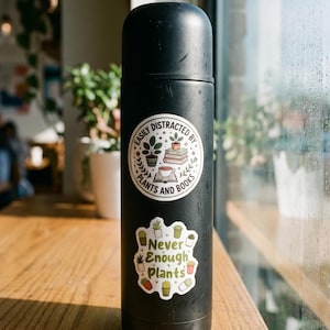 May include: A matte black cylindrical insulated water bottle featuring two circular stickers. The top sticker reads EASILY DISTRACTED BY PLANTS AND BOOKS with illustrations of potted plants, books, and a cup. The bottom sticker says Never Enough Plants.
