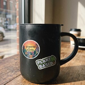May include: A black coffee mug with two stickers on it. One sticker says "powered plants" with a floral design, and the other says "PLANT BASED" with a green leaf. The mug sits on a wooden table.