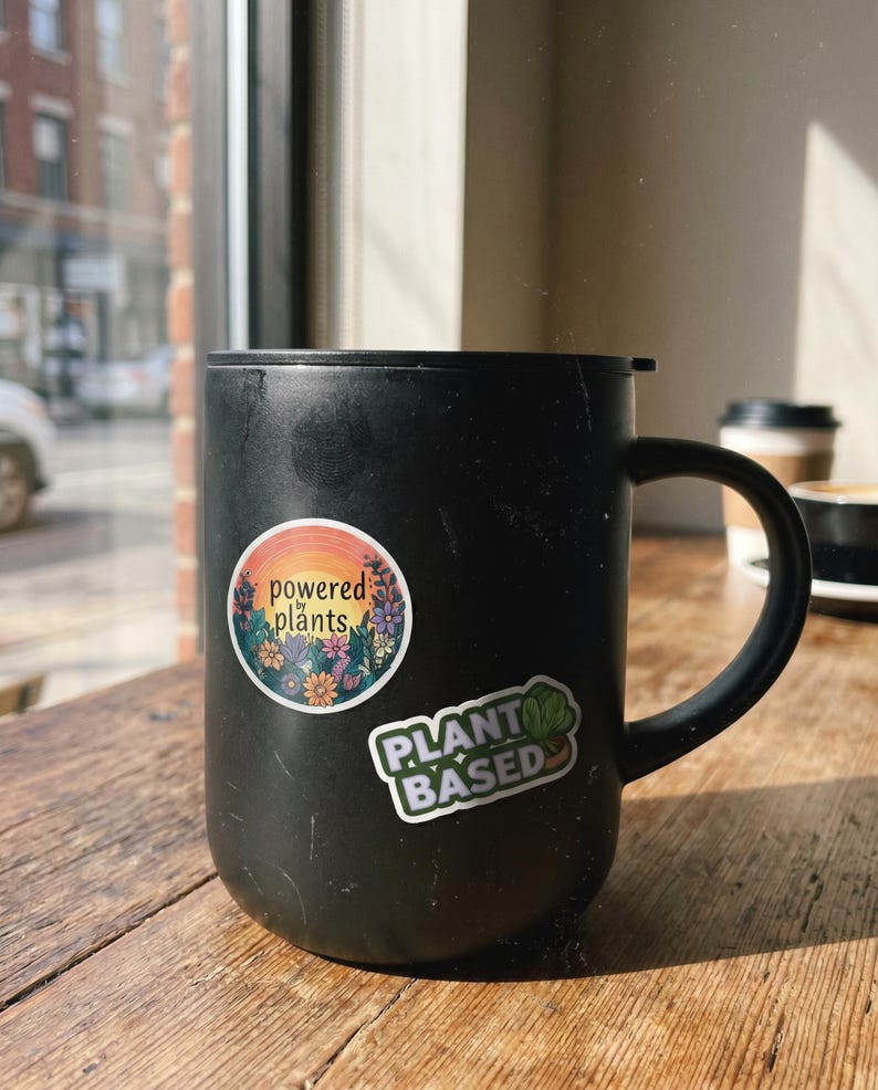 May include: A black coffee mug with two stickers on it. One sticker says "powered plants" with a floral design, and the other says "PLANT BASED" with a green leaf. The mug sits on a wooden table.